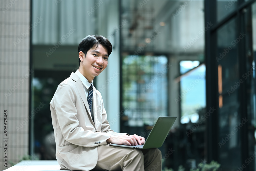 Smiling young businessman working on laptop outside modern office building Smiling young businessman working on laptop outside modern office building
