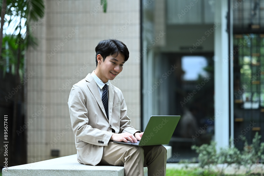 Attractive businessman in suit sitting on bench and checking email on laptop before meeting Attractive businessman in suit sitting on bench and checking email on laptop before meeting