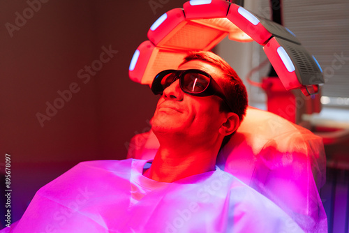 Male patient with hairloss problem in protective goggles having LED light hair photodynamic therapy treatment after hair transplant at clinic. Man client having non-invasive type of phototherapy. photo