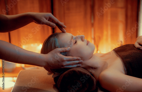 Caucasian woman enjoying relaxing anti-stress head massage and pampering facial beauty skin recreation leisure in warm candle lighting ambient salon spa in luxury resort or hotel. Quiescent