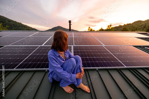 Fotografía Rooftop with solar panels on house in mountains, woman sitting alone raising hands and enjoying sunset