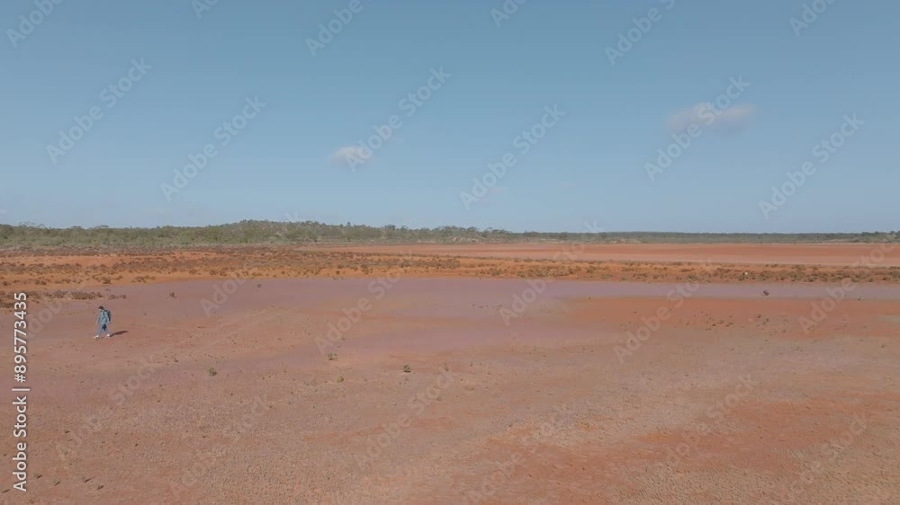 Panning drone clip from right to left, showing rugged Australian outback habitat and vegetation, and male backpacker walking on baked orange desert earth Panning drone clip from right to left, showing rugged Australian outback habitat and vegetation, and male backpacker walking on baked orange desert earth