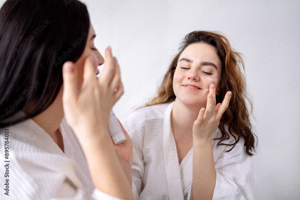 Young women in bathrobes apply cream on their faces at the spa. Charming friends take care of their skin in a beauty salon. Young women in bathrobes apply cream on their faces at the spa. Charming friends take care of their skin in a beauty salon.
