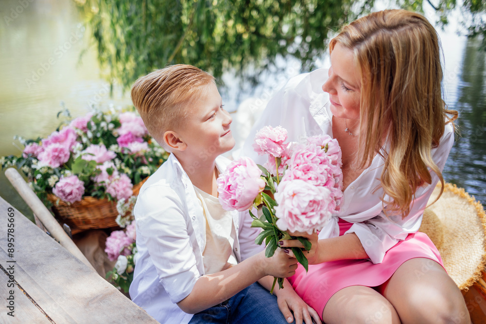 Cute boy and his attractive mother on a wooden boat on a pond or lake. A smiling son gives his blonde mom pink peonies. Cute boy and his attractive mother on a wooden boat on a pond or lake. A smiling son gives his blonde mom pink peonies.