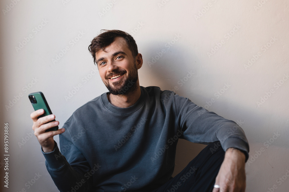 Handsome happy man sitting on the floor, hold green mobile phone, wear grey pullover, black pants. Smiling man chatting, messaging or read news in his mobile device, he is laughing. Handsome happy man sitting on the floor, hold green mobile phone, wear grey pullover, black pants. Smiling man chatting, messaging or read news in his mobile device, he is laughing.