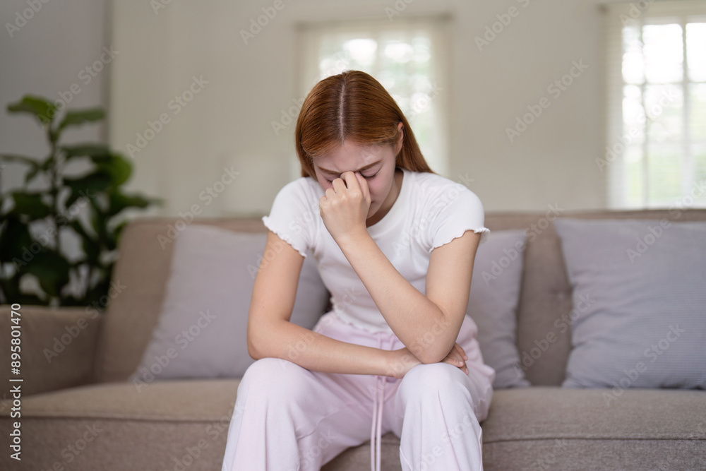 Young Woman Sitting on Couch Holding Head in Pain, Experiencing a Severe Headache or Migraine at Home Young Woman Sitting on Couch Holding Head in Pain, Experiencing a Severe Headache or Migraine at Home