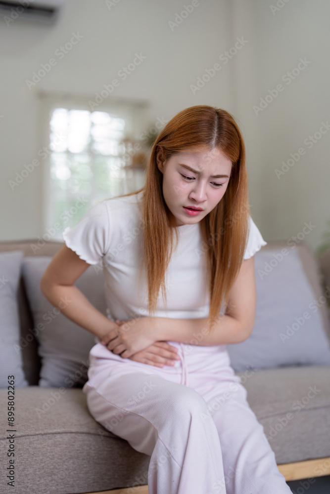 Young Woman Experiencing Stomachache While Sitting on a Couch at Home, Holding Her Abdomen in Pain Young Woman Experiencing Stomachache While Sitting on a Couch at Home, Holding Her Abdomen in Pain