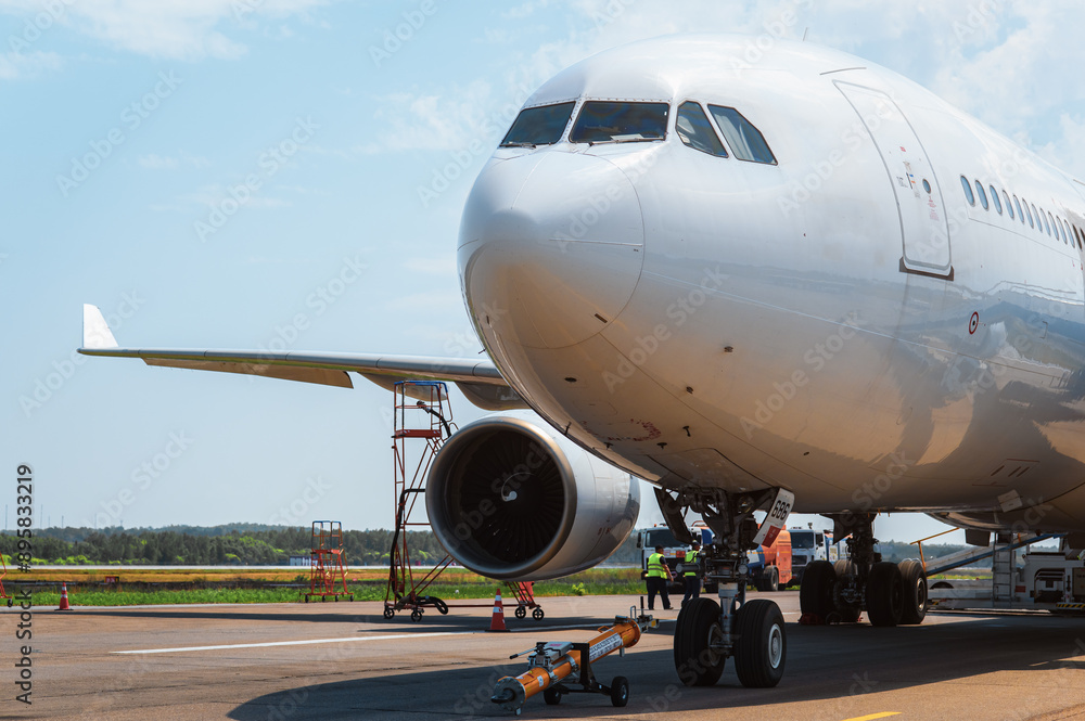 A wide-body passenger jet airliner undergoes pre-takeoff servicing and fueling on the apron on a sunny day. The cockpit and fuselage of the aircraft reflect the runway and blue sky A wide-body passenger jet airliner undergoes pre-takeoff servicing and fueling on the apron on a sunny day. The cockpit and fuselage of the aircraft reflect the runway and blue sky