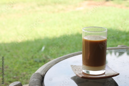 A glass of wedang kopi jahe or hot ginger coffee sitting on a table. Traditional Indonesian hot beverage. Blurry green park in the background. Selective focus. photo