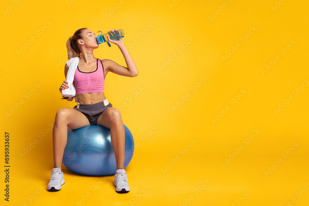 Sporty Woman Sitting On Fitball Drinking Water Over Pink Background In Studio. Workout Break Concept. Empty Space Sporty Woman Sitting On Fitball Drinking Water Over Pink Background In Studio. Workout Break Concept. Empty Space