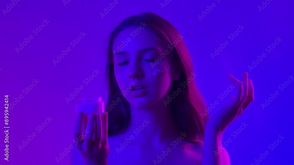 Young woman posing at camera holds jar applies cosmetic product on skin, isolated on blue neon background. Natural beauty skin care concept Young woman posing at camera holds jar applies cosmetic product on skin, isolated on blue neon background. Natural beauty skin care concept