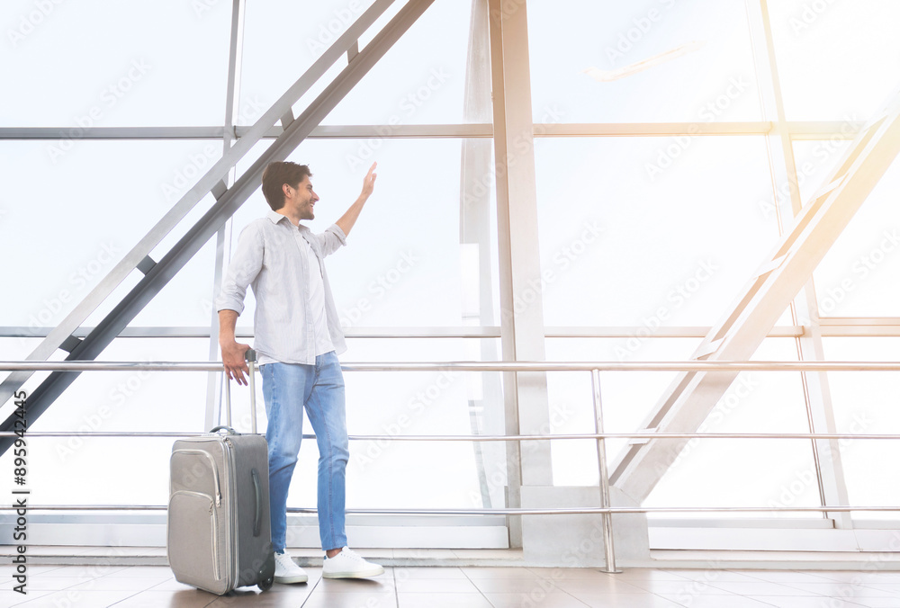 Welcoming New City. Happy Man Traveler Walking With Luggage In Airport, And Waving To Window, Excited About Trip, Free Space Welcoming New City. Happy Man Traveler Walking With Luggage In Airport, And Waving To Window, Excited About Trip, Free Space
