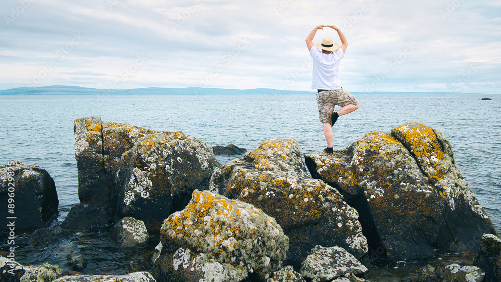 Handsome bearded man with straw hat doing yoga on the rocks by the ocean at wild Atlantic way, adventure and lifestyle concept, summer vacation, nature background