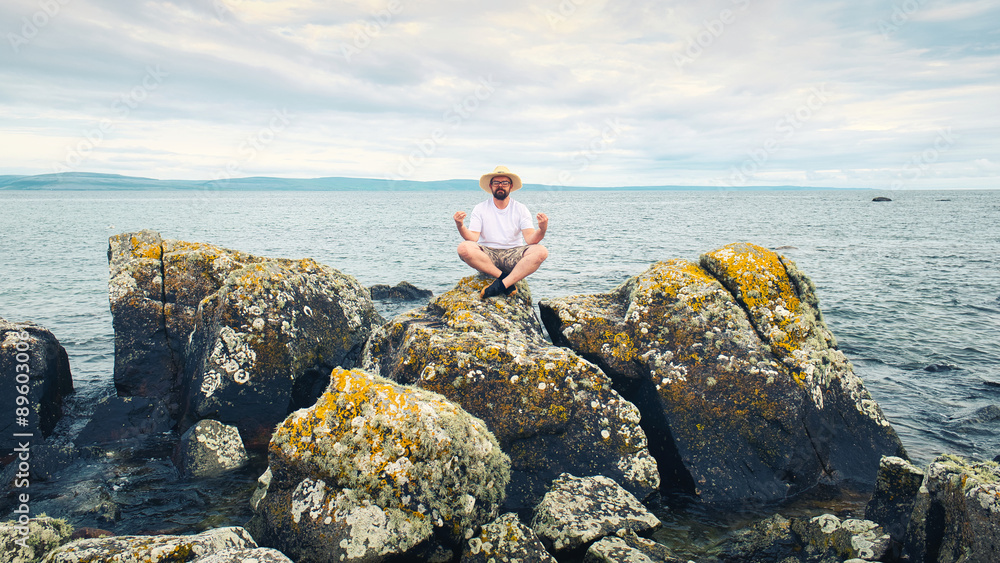 Handsome bearded man with straw hat doing yoga on the rocks by the ocean at wild Atlantic way, adventure and lifestyle concept, summer vacation, nature background