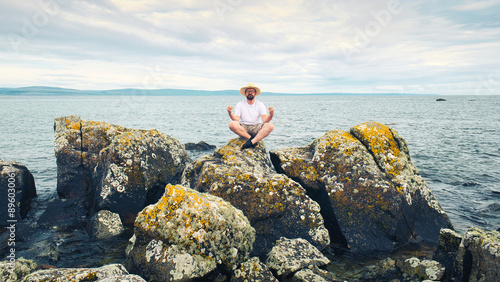 Obraz na płótnie Handsome bearded man with straw hat doing yoga on the rocks by the ocean at wild Atlantic way, adventure and lifestyle concept, summer vacation, nature background w salonie