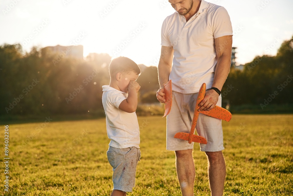 Broken toy plane, boy is crying. Father and little son are together on the summer field Broken toy plane, boy is crying. Father and little son are together on the summer field
