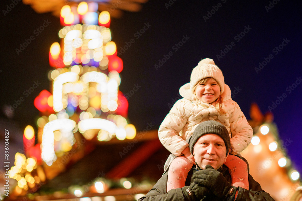Little preschool girl sitting on shoulder of father on Christmas market in Germany. Happy toddler child and man observing traditional decorated pyramid. Happy family, bonding, love. Family xmas time. Little preschool girl sitting on shoulder of father on Christmas market in Germany. Happy toddler child and man observing traditional decorated pyramid. Happy family, bonding, love. Family xmas time.