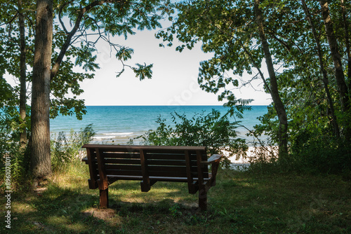 Fototapeta Naklejka Na Ścianę i Meble -  A shaded park bench affords a visitor an overlooking view of the calm waters of Lake Michigan from Harrington Beach State Park, Belgium, Wisconsin in early July.