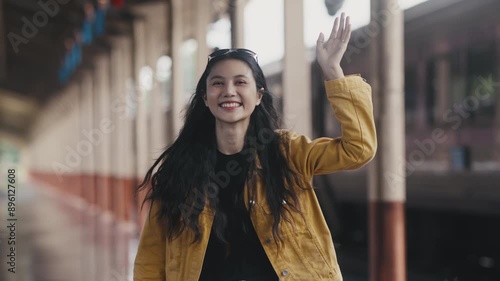 Young woman warmly greets onlookers at a quiet train station platform. She waves and smiles, radiating happiness and excitement