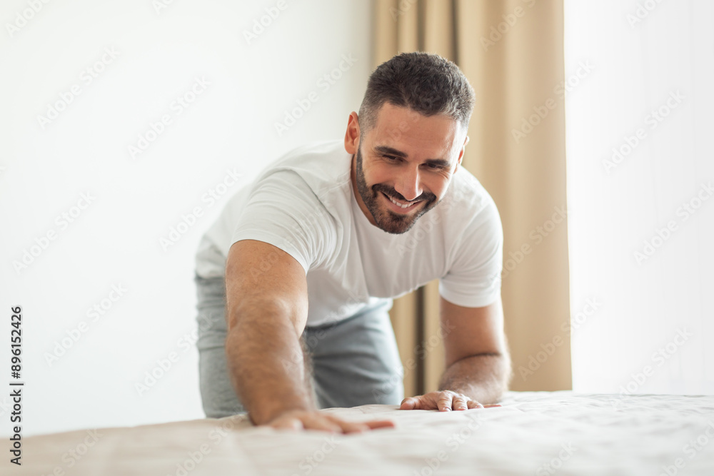 A man in a white shirt and gray pants is bending over and making a bed. He is smiling and looks happy as he smooths the bedspread. The room is bright and airy, with a large window in the background. A man in a white shirt and gray pants is bending over and making a bed. He is smiling and looks happy as he smooths the bedspread. The room is bright and airy, with a large window in the background.