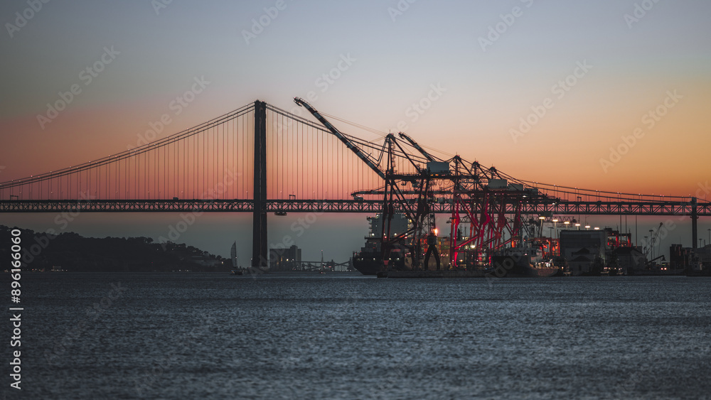 Fototapeta premium A panoramic view of a shipping port with cranes and a cargo ship against the backdrop of a bridge during sunset. The sky with vibrant colors creating a stunning silhouette of the industrial structures