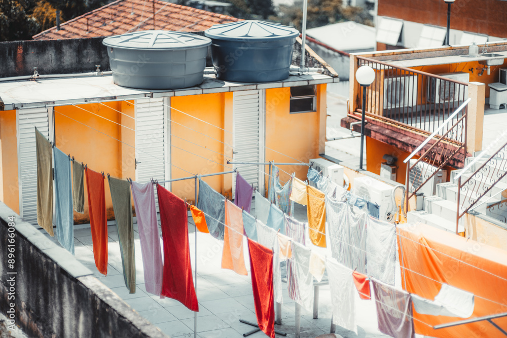 Clothes and towels hanging to dry on a rooftop laundry area of a building. The space features bright orange and yellow walls, white shutters, water tanks on the roof, and outdoor staircases Clothes and towels hanging to dry on a rooftop laundry area of a building. The space features bright orange and yellow walls, white shutters, water tanks on the roof, and outdoor staircases