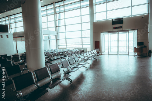 A spacious, empty airport waiting area with rows of metal chairs arranged neatly. Large floor-to-ceiling windows flood the space with natural light, creating a bright, airy atmosphere photo