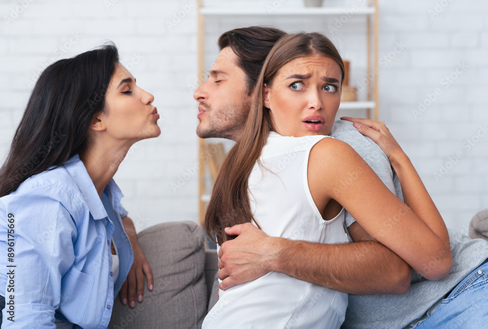 Jealousy And Infidelity. Girl Hugging Cheating Boyfriend While He Kisses Her Friend Sitting Together On Sofa At Home Jealousy And Infidelity. Girl Hugging Cheating Boyfriend While He Kisses Her Friend Sitting Together On Sofa At Home