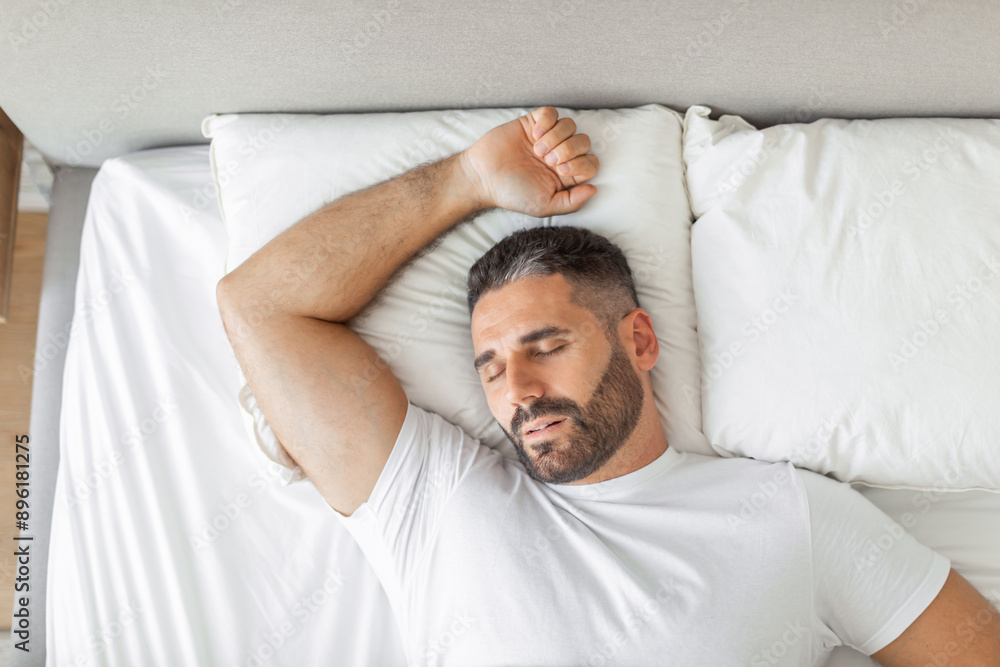 A man with a beard is sleeping soundly in a white bed. His arm is extended above his head, resting on a pillow. The man is wearing a white shirt and his eyes are closed. A man with a beard is sleeping soundly in a white bed. His arm is extended above his head, resting on a pillow. The man is wearing a white shirt and his eyes are closed.