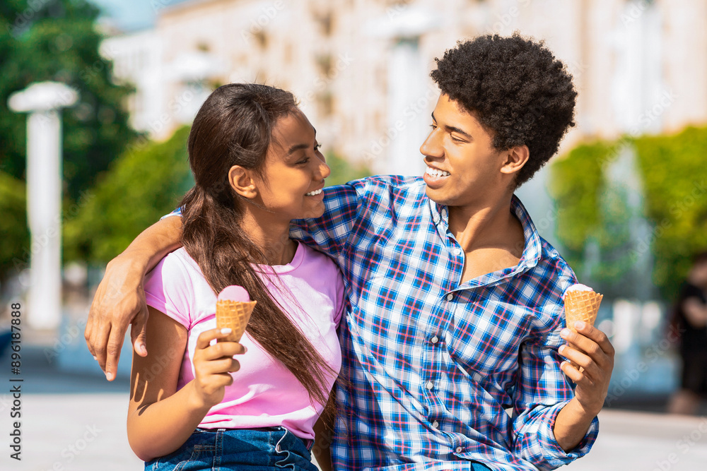 Fototapeta premium A teen black couple sitting outside in a city setting, both holding ice cream cones. They are looking at each other and smiling, enjoying their treat and each others company on a warm, sunny day.