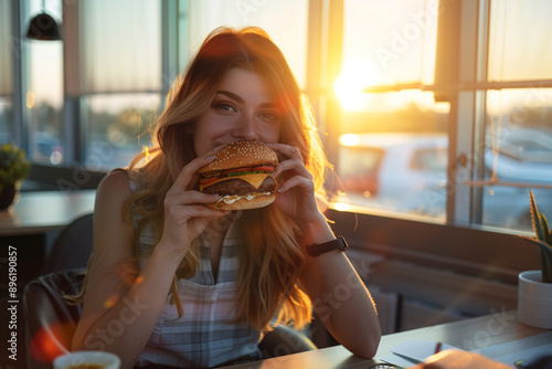 Beautiful girl eating burger in cafe. Looking straight into camera. Setting sun on background. photo