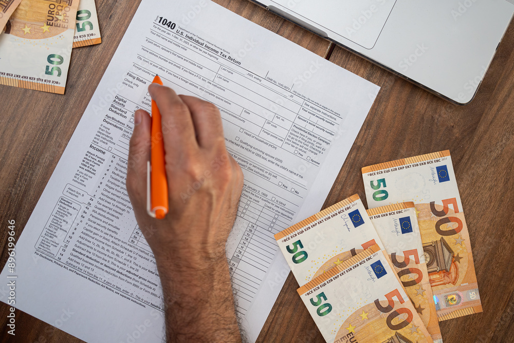 Obraz premium Trader carefully fills 1040 form with pen surrounded by euro bills and laptop at table reporting gains and losses from digital asset transactions to calculate tax