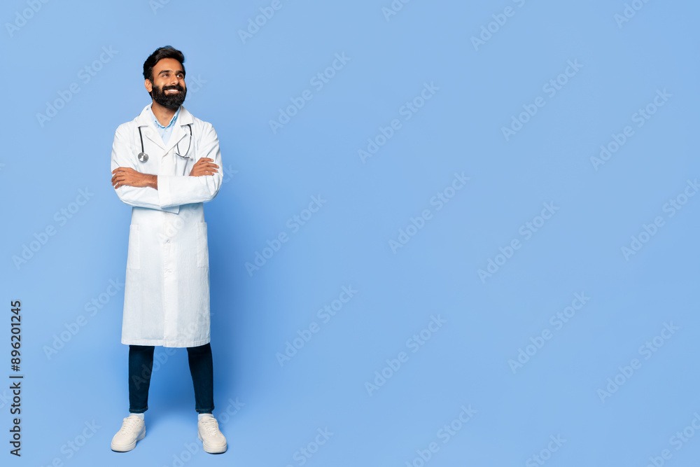 A bearded Indian man doctor, wearing a white lab coat, stands with his arms crossed against a blue background. He is looking at copy space, with a slight smile on his face A bearded Indian man doctor, wearing a white lab coat, stands with his arms crossed against a blue background. He is looking at copy space, with a slight smile on his face