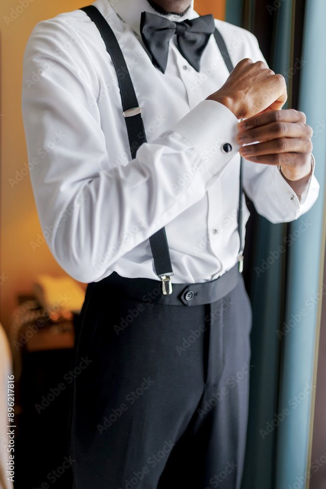 A dark-skinned man puts on a white shirt and buttons the cuff close-up. Stylish groom puts on a white shirt and suspenders while preparing for the wedding ceremony A dark-skinned man puts on a white shirt and buttons the cuff close-up. Stylish groom puts on a white shirt and suspenders while preparing for the wedding ceremony