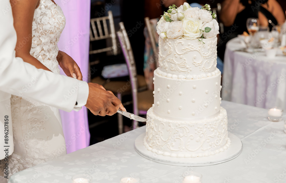 The bride and groom cut a white wedding cake at a banquet in a restaurant. Hands of the bride and groom close up holding one knife together. The ceremony of cutting the cake for dark skinned newlyweds The bride and groom cut a white wedding cake at a banquet in a restaurant. Hands of the bride and groom close up holding one knife together. The ceremony of cutting the cake for dark skinned newlyweds