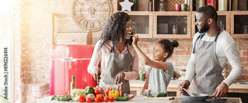 Little black girl feeding mom while cooking together with parents at home, panorama