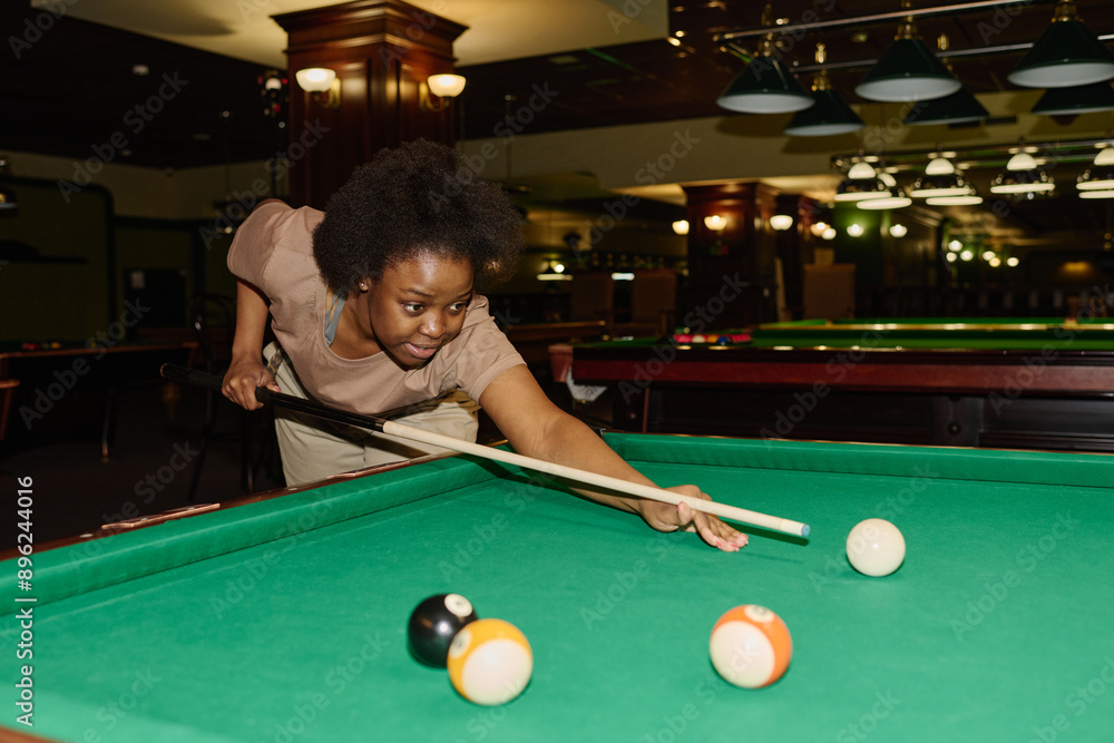 Fototapeta premium Young active woman in casual attire bending over pool table and looking at one of balls before hitting it with cue while playing billiards