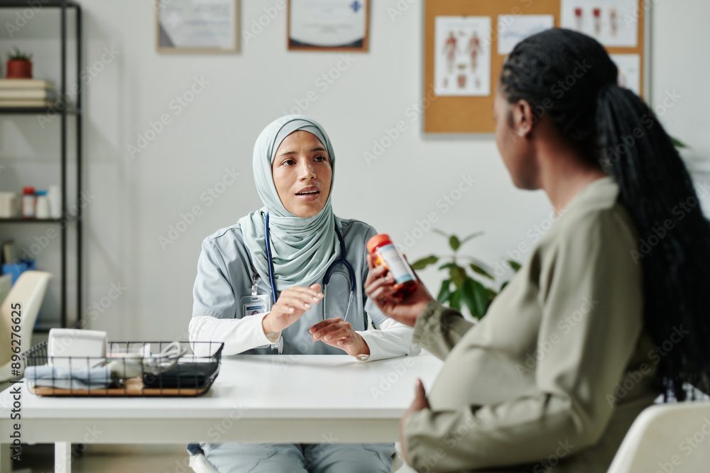 Young confident clinician in hijab and medical scrubs sitting by desk in front of pregnant patient and consulting her about new pills Young confident clinician in hijab and medical scrubs sitting by desk in front of pregnant patient and consulting her about new pills