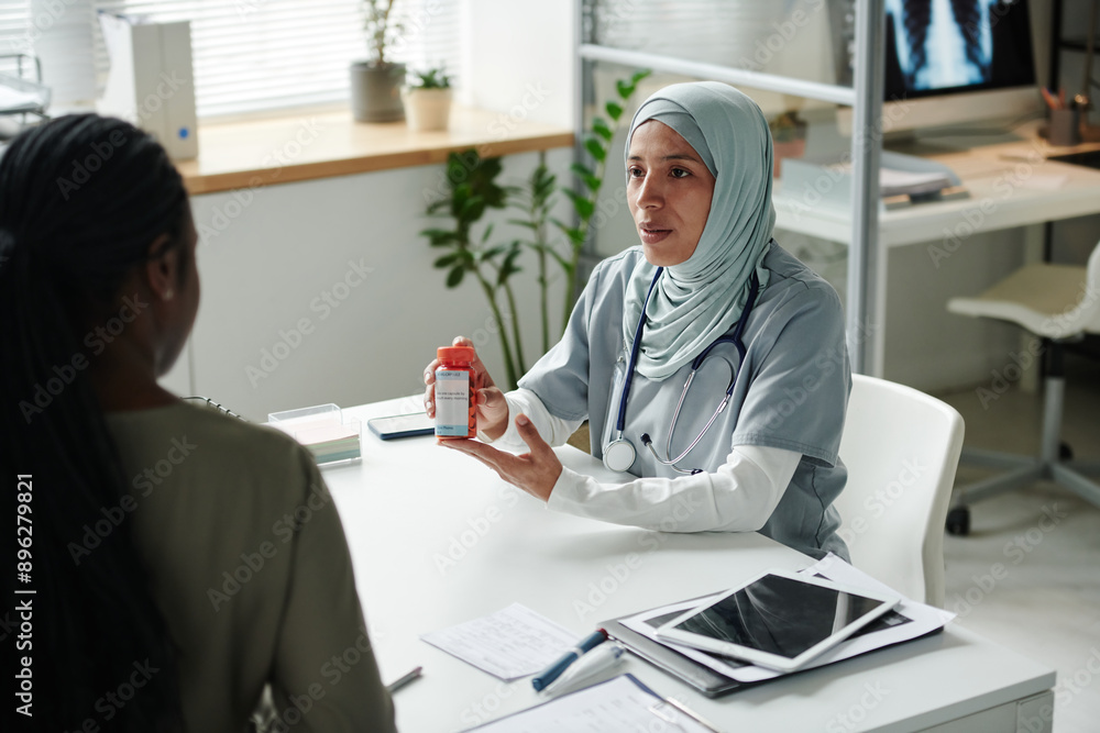 Obraz premium Young confident female clinician in medical scrubs and hijab prescribing pills to African American patient and describing its characteristics