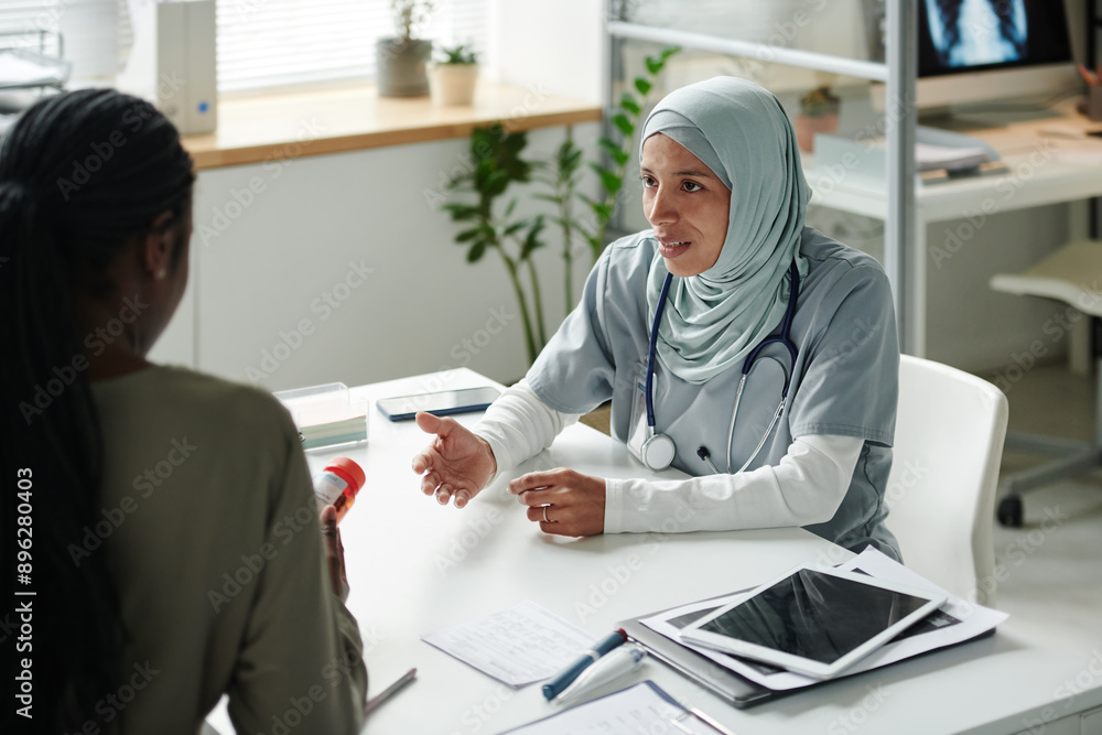 Obraz premium Young Hispanic woman in hijab and medical scrubs looking at female patient with bottle of pills or vitamins during consultation