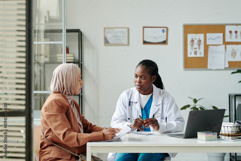 Obraz premium Young African American female clinician in lab coat talking to Muslim patient in hijab and casual apparel during medical consultation