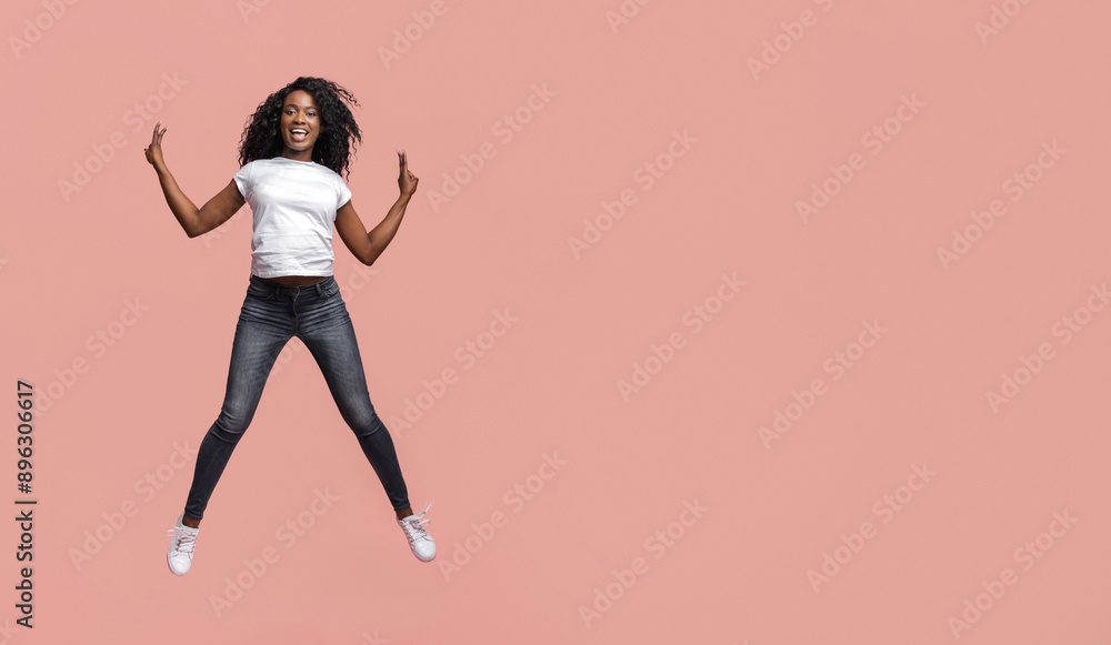 This image shows a young African American woman with curly hair jumping in front of a plain pink background. She is wearing a white t-shirt, dark blue jeans, and white sneakers. This image shows a young African American woman with curly hair jumping in front of a plain pink background. She is wearing a white t-shirt, dark blue jeans, and white sneakers.