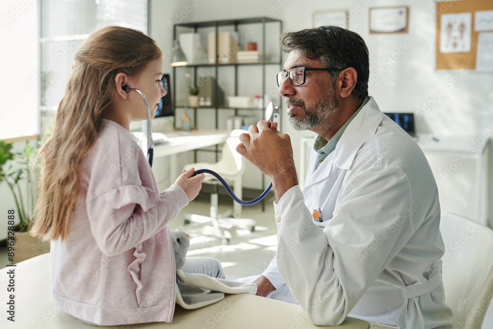 Obraz premium Cute little girl with stethoscope playing doctor with mature male pediatrician while visiting him in modern clinics