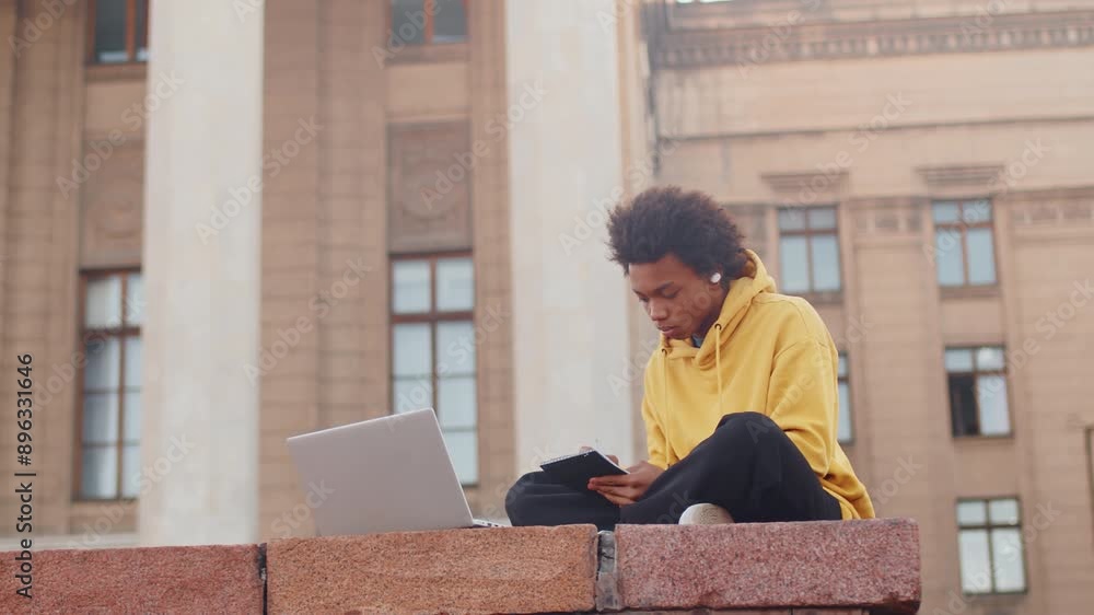 Young black programmer in casual clothes sitting with a laptop outdoors near the building and looking at a smartphone. Concept of flexible remote work Young black programmer in casual clothes sitting with a laptop outdoors near the building and looking at a smartphone. Concept of flexible remote work