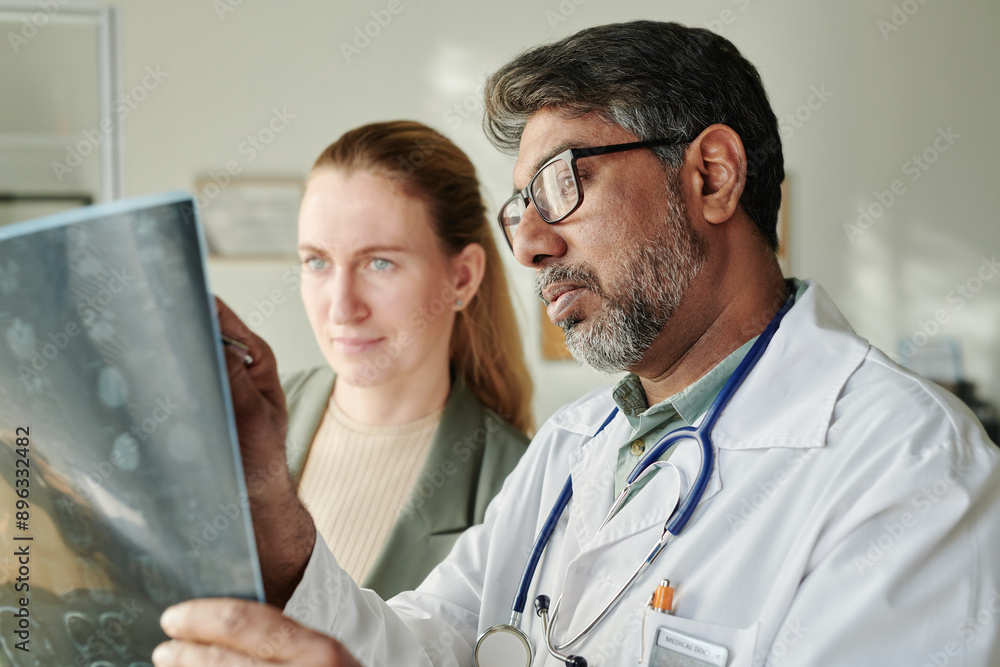 Confident male clinician in eyeglasses standing next to young female patient in medical office and commenting her x-ray image Confident male clinician in eyeglasses standing next to young female patient in medical office and commenting her x-ray image