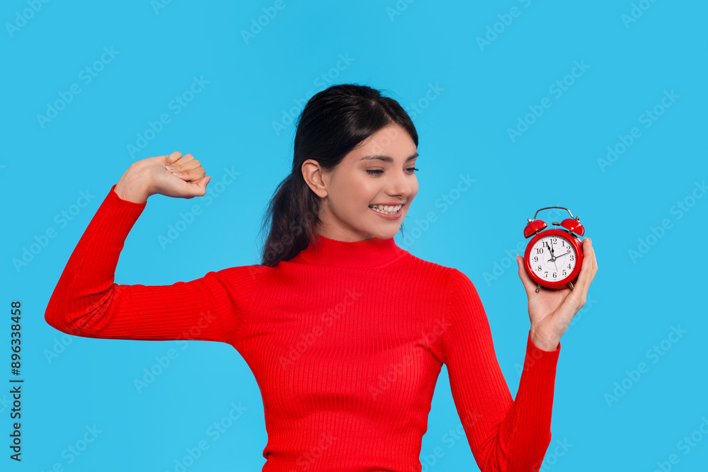 Smiling Woman In Red Flexes Bicep While Holding Red Alarm Clock Against Blue Background Smiling Woman In Red Flexes Bicep While Holding Red Alarm Clock Against Blue Background