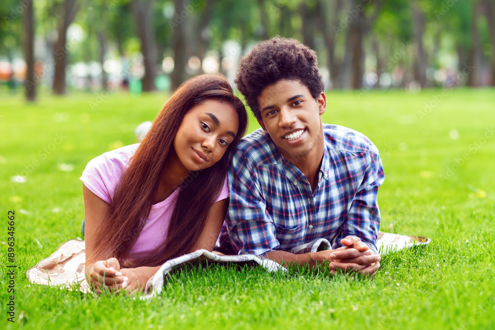 A teen black couple smiles at the camera while lying on a blanket in a grassy park. The girl is resting her head on guys shoulder. They are enjoying a sunny summer day together. A teen black couple smiles at the camera while lying on a blanket in a grassy park. The girl is resting her head on guys shoulder. They are enjoying a sunny summer day together.