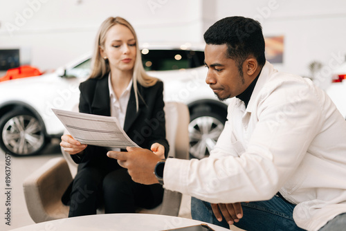 Medium shot of female manager consulting doubtful black buyer, giving purchase agreement for review at showroom. Thoughtful African client receiving car purchase contract before signing in dealership photo