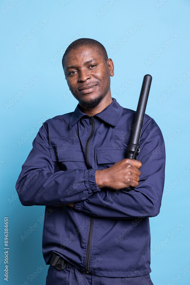 African american security guard feels proud with his police weapon on camera, posing with confidence with his baton and professional uniform. Trained bodyguard against blue background. African american security guard feels proud with his police weapon on camera, posing with confidence with his baton and professional uniform. Trained bodyguard against blue background.