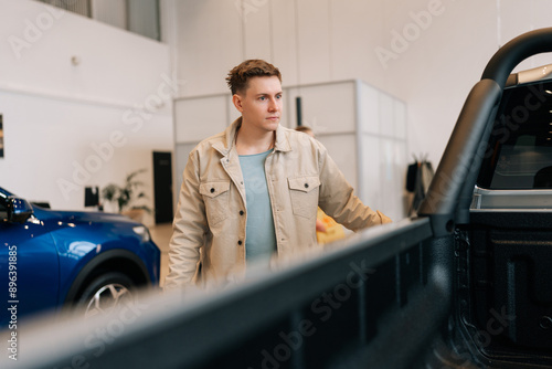 Young man buyer looking around big open trunk of car. Male customer choosing pickup truck in car dealership. Guy visitor walking in modern auto showroom and choosing new vehicle to buy or rent.